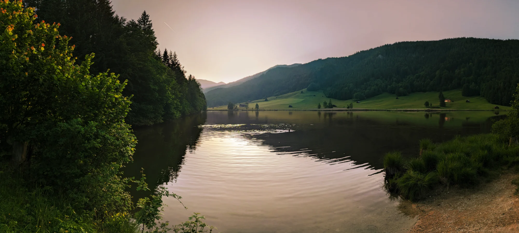 Sonnenuntergang über dem Geroldsee mit ruhigem Wasser, umrahmt von Wiesen, Bäumen und sanften Hügeln.