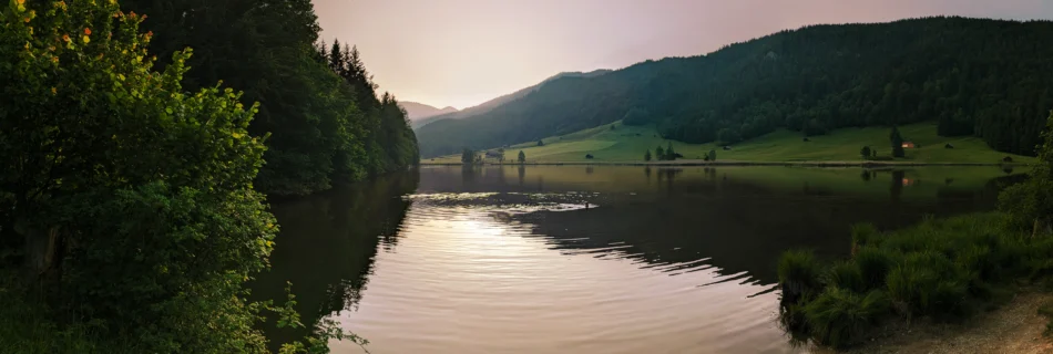Sonnenuntergang über dem Geroldsee mit ruhigem Wasser, umrahmt von Wiesen, Bäumen und sanften Hügeln.