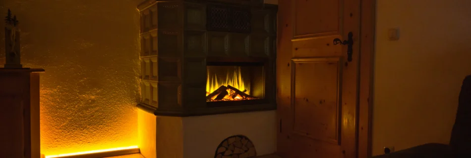 Wohnzimmer im Ferienhaus Wiesenhof mit traditionellem Kachelofen und integriertem Elektrokamin – stimmungsvolle Beleuchtung und warme Atmosphäre.